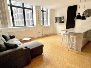 Ein modernes Wohnzimmer mit einem grauen Sofa, einem kleinen runden Tisch, einem weißen Barhocker, einem Fernseher und einem großen Fenster.