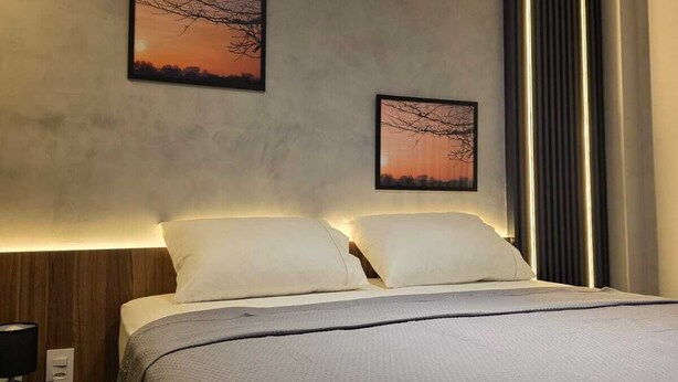 Una habitación de hotel moderna con una cama grande, dos obras de arte colgadas en la pared que representan puestas de sol, y una mesita de noche con una lámpara.