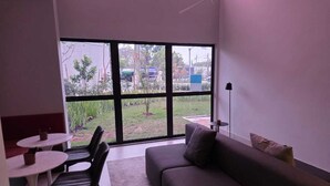 Una habitación moderna con un sofá gris, dos sillas negras, una mesa redonda pequeña y un ventanal que ofrece vistas a un jardín y una calle.