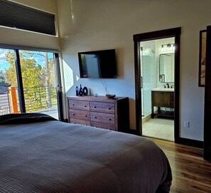 A bedroom with a large bed, a wooden dresser, a flat-screen TV, and a bathroom visible through an open door. - The Riverlight 3 Bedroom Townhouse (Helena)