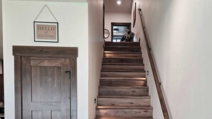 Eine Holztreppe mit Handlauf, eine Tür mit der Aufschrift 'HELLO AND WELCOME' und ein Lichtschalter an der Wand.