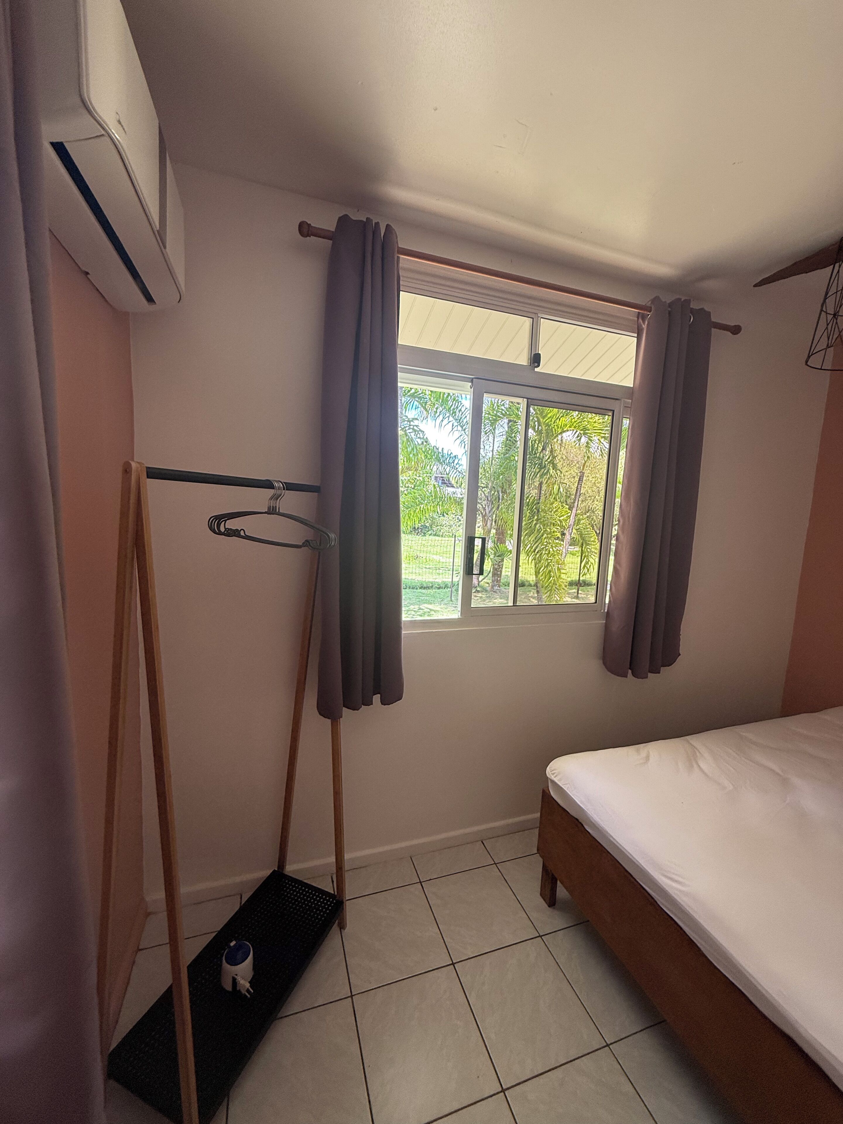 Una habitación pequeña y limpia con una cama, un ventanal con cortinas, un radiador y una mesita con hervidor.