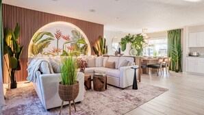 A modern living room with a sectional sofa, a coffee table, and a dining area with a wooden table and chairs. There are various potted plants and a large window providing natural light. - The Don | Fully Loaded Waterfront Wellness (Tampa)