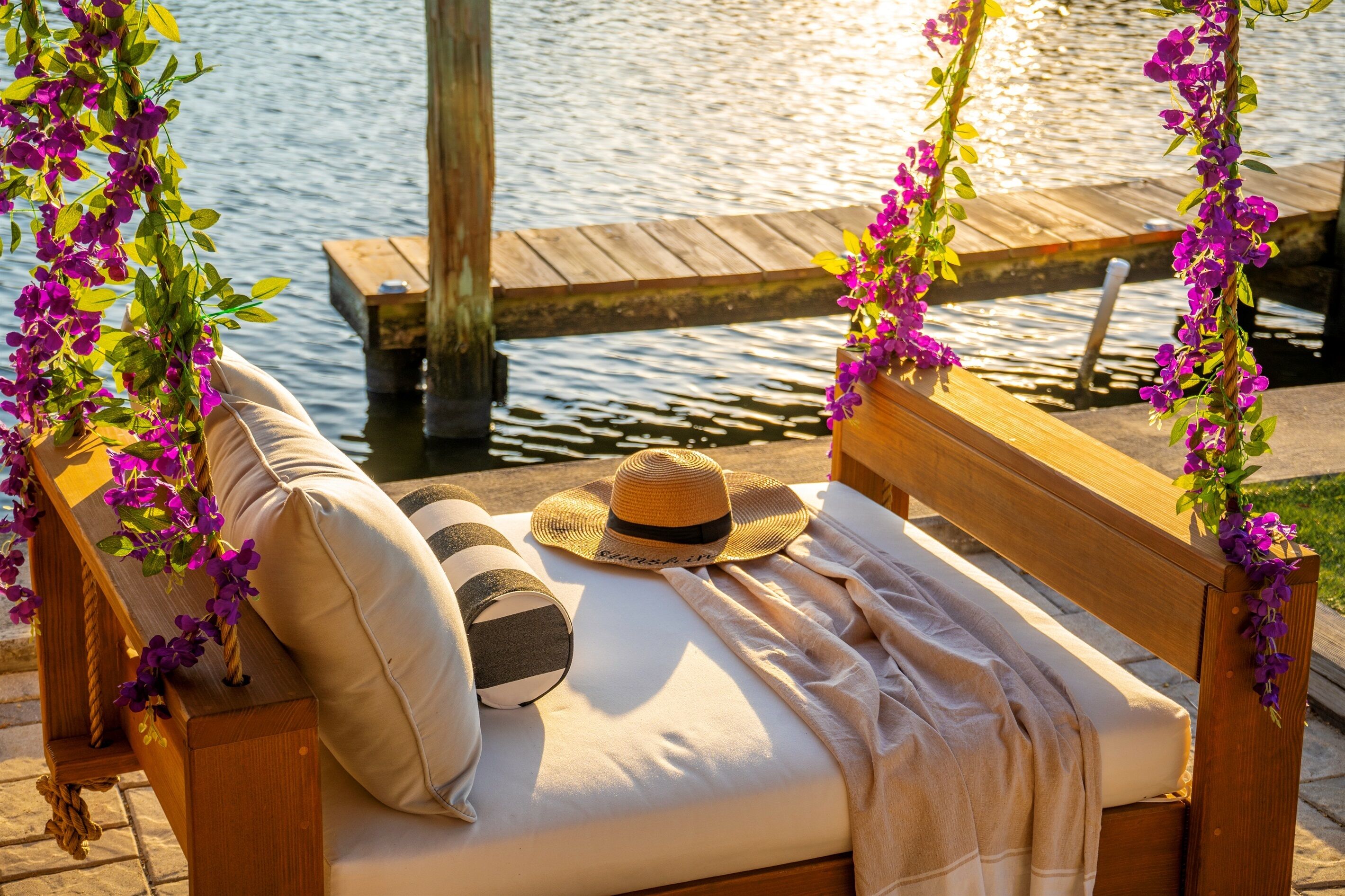 A wooden bench with a cushion and a hat, surrounded by purple flowers, overlooking a wooden dock and water.