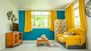 A room with a teal wall, yellow curtains, a wooden cabinet, a sofa, and a small table. - Vahine Ori Lodge (Tumaraa)