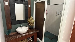 Un baño con tocador de madera, un lavamanos blanco, un espejo, zona de ducha con puerta de vidrio y azulejos verdes.