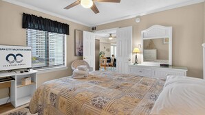 A bedroom with a bed, a desk, a chair, a ceiling fan, a window with blinds, and a mirror. - 9400 Building #401 (Ocean City)