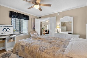 Ein Schlafzimmer mit Bett, Schreibtisch, Stuhl, Deckenventilator, Fenster mit Jalousien und Spiegel.