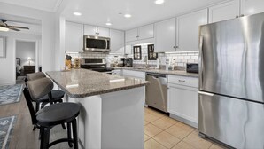 A modern kitchen with white cabinets, stainless steel appliances, and a granite countertop island with bar stools. - 9400 Building #401 (Ocean City)