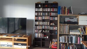 A living room with a large flat-screen TV, a bookshelf filled with books and DVDs, and a small table with a few items on it. - 3B parkview Sancatury Lake house for 6people &Wifi&Lake&golf&SPA (Point Cook)