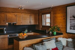 A modern kitchen with wooden cabinetry, a central island with a fruit bowl, and a cozy seating area with a sofa and cushions. - Luxury Ski Apartment Valentine in Verbier, Switzerland LVRB-4 (Verbier)