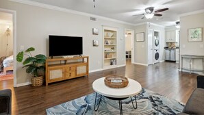 A spacious living room with hardwood floors, a flat-screen TV, a round coffee table, and a ceiling fan.
