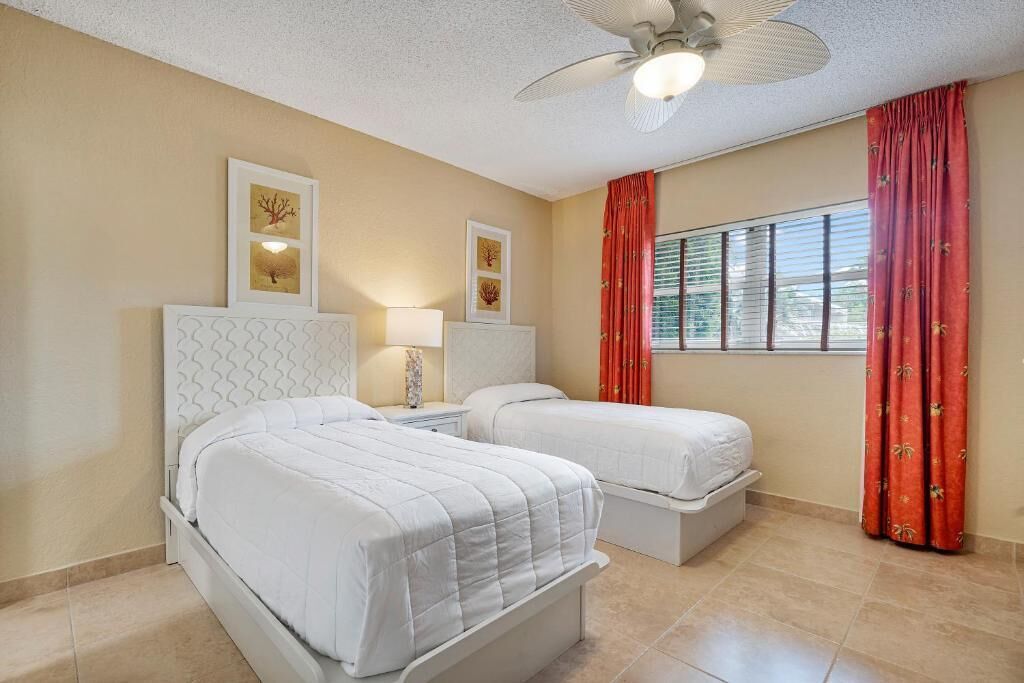 Une chambre d’hôtel avec deux lits, un ventilateur de plafond, une fenêtre avec des rideaux rouges et des œuvres d’art accrochées aux murs.