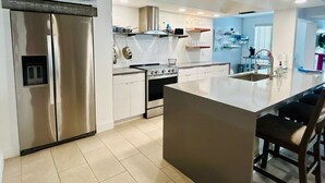 A modern kitchen with stainless steel appliances, a central island, and a dining area. - Pink Pineapple ~ Walk to Downtown ~ Chic Heated Pool Home (Cape Coral)
