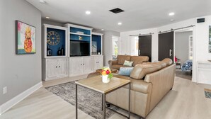 A modern living room with a beige leather sofa, a wooden coffee table with a vase of flowers, and a large blue cabinet with a clock. - Pink Pineapple ~ Walk to Downtown ~ Chic Heated Pool Home (Cape Coral)