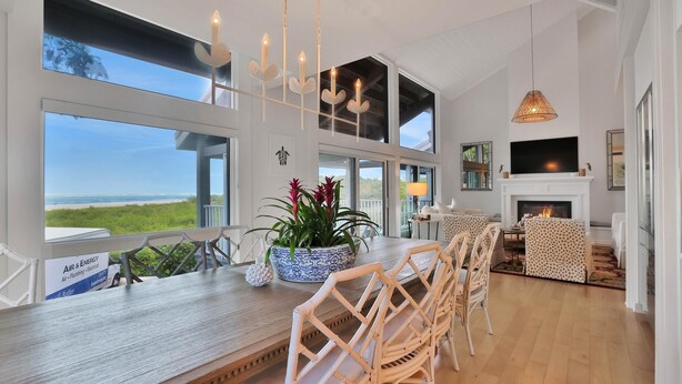 Un vaste salon doté d’une grande fenêtre donnant sur la plage, d’une cheminée et d’un coin repas avec des chaises en osier et une table en bois.