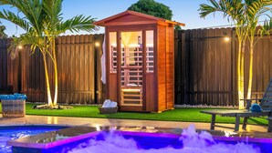 A wooden sauna structure with a glass door, a small pool with a fountain, and a wooden fence in the background.