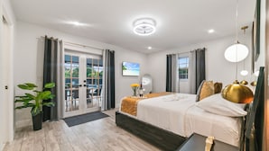 A modern hotel room with a large bed, a bedside table, a potted plant, and a view of the outdoors through the sliding glass door.