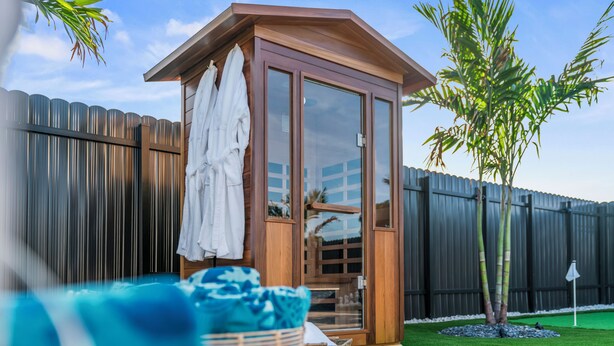 A wooden cabin with a glass door, a white towel hanging on the door, and a small garden area with a tree and artificial grass.