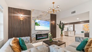 A modern living room with a large flat-screen TV, tufted sofas, a coffee table, and a kitchen in the background.