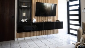 A modern living room with a flat-screen TV mounted on a wooden panel wall, a dark wooden cabinet below, and a large window with blinds. - Rubi Ocean House (Cabo Rojo)