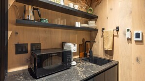 Eine kompakte Küche mit einer schwarzen Mikrowelle, einer Kaffeemaschine und einem Spülbecken. Darüber befinden sich Holzregale, auf denen Tassen und eine Vase mit Trockenblumen stehen.