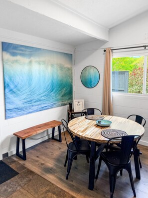 A dining area with a round wooden table, black chairs, a bench, and a large wave painting on the wall. - Beachy Bungalow (Waialua)
