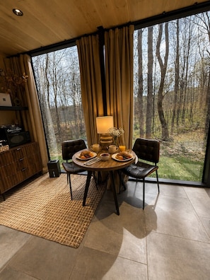 A dining area with a table set for two, a lamp, and a view of a forest through large windows.