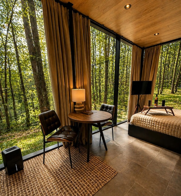 A modern room with a large window, a round table, two chairs, a bed, and a television.