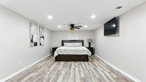 Un dormitorio con suelo de madera, una cama con cubrecama blanco, ventilador de techo, iluminación empotrada y obras de arte en las paredes.