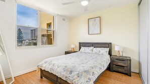 A bedroom with a bed, two bedside tables, a ceiling fan, a window with a view, and a painting on the wall. - Oceanfront Stay Steps to the Sand (The Entrance)