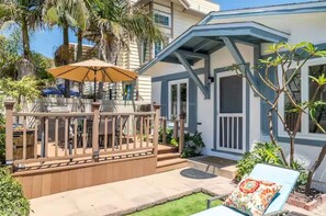A house with a wooden deck, a covered patio, and a small garden area with a chair and umbrella. - Roswitha's Blue Wave Cottage (San Diego)