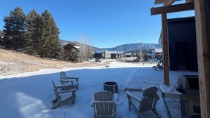 Un patio cubierto de nieve con sillas Adirondack y una parrilla, con vistas a un paisaje montañoso.