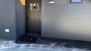 Un edificio con fachada gris, un porche de madera, una puerta y un ventanal pequeño.