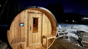 Estructura de sauna de madera con puerta y una ventana pequeña, rodeada de nieve y algunas sillas.