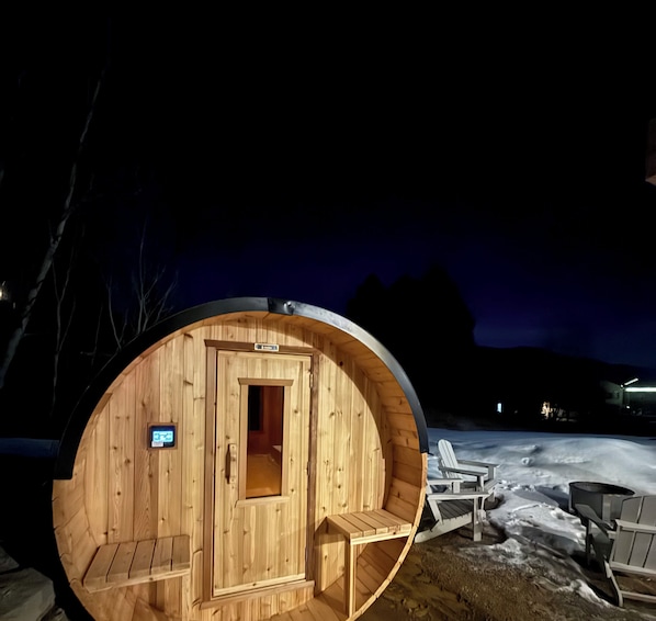 Une structure de sauna en bois, dotée d’une porte et d’une petite fenêtre, entourée de neige et de quelques chaises.