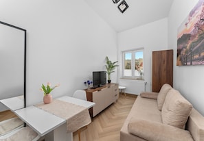 A modern living room with a white sofa, a TV on a wooden cabinet, a dining table with a plant, and a large window with a view. - Aleja 29 Listopada Cozy Studio (Kraków)