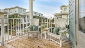 A covered porch with white Adirondack chairs and a small table, overlooking a residential area with multiple houses. - New! Coastal House, Steps to Beach, Pool, Great Location! (Gulf Shores)