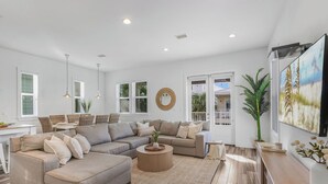 A spacious living room with a large sectional sofa, a round coffee table, and a dining area with a white table and chairs. - New! Coastal House, Steps to Beach, Pool, Great Location! (Gulf Shores)