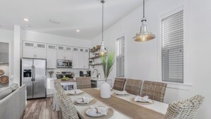A modern kitchen with white cabinetry, a stainless steel refrigerator, and a dining area with a white table set for four. - New! Coastal House, Steps to Beach, Pool, Great Location! (Gulf Shores)