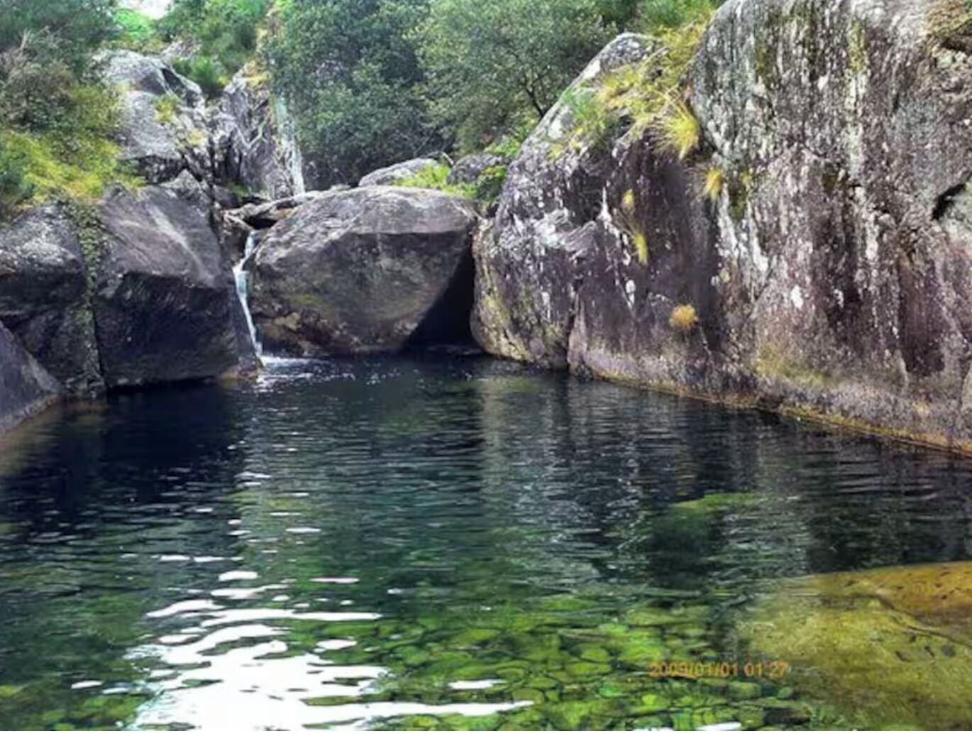 A natural landscape with a waterfall, large rocks, and a clear water body surrounded by greenery.