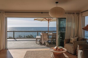 A room with a view of the sea, a wooden deck, a table with chairs, a sofa, and a hanging light fixture. - Waterview Wombarra (Wombarra)
