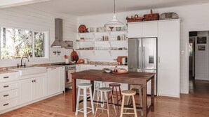 Una cocina con armarios blancos, una isla de madera con taburetes, un refrigerador de acero inoxidable y una ventana con vistas al verdor.