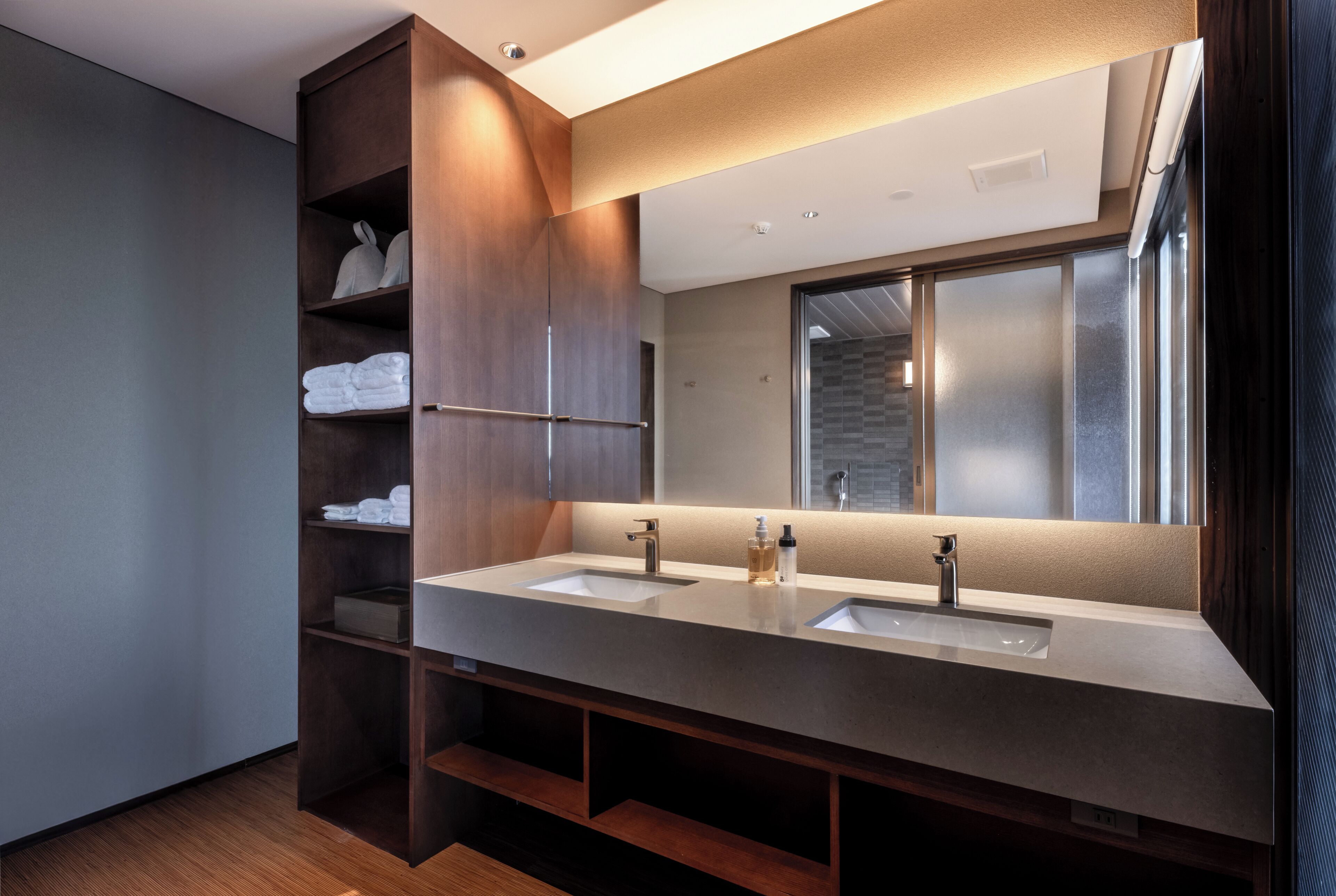 A modern bathroom with two sinks, a large mirror, and wooden shelving with towels.