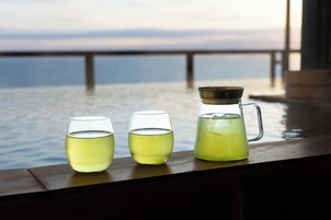 Una mesa con tres vasos y una jarrón de líquido amarillo, frente a una vista del agua y una barandilla de madera.