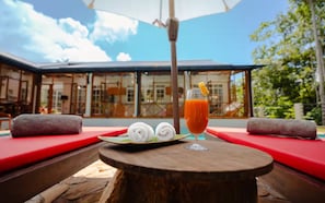 A table with a glass of orange juice, two rolled towels, and a plate with a slice of lemon, set against a backdrop of a building with large windows and a clear sky. - Sevena Villa: Magical Home with a Yoga Shala (Weligama)
