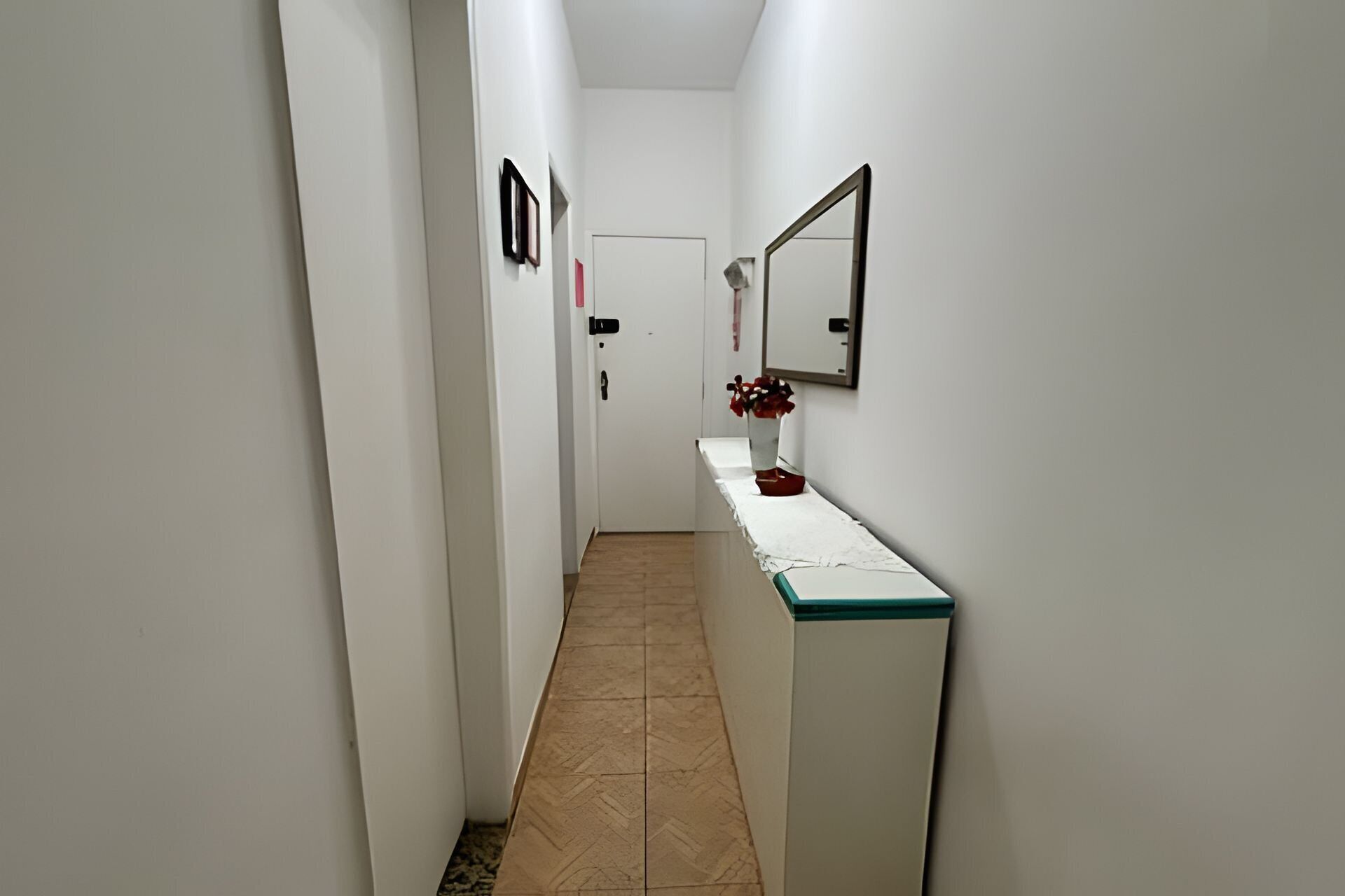 A narrow hallway with white walls, a tiled floor, a door at the end, and a counter with a mirror and a vase of flowers.