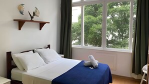 Ein ordentlich bezogenes Bett mit weißer und blauer Bettwäsche, ein Holz-Kopfteil und ein Fenster mit Blick auf Bäume.