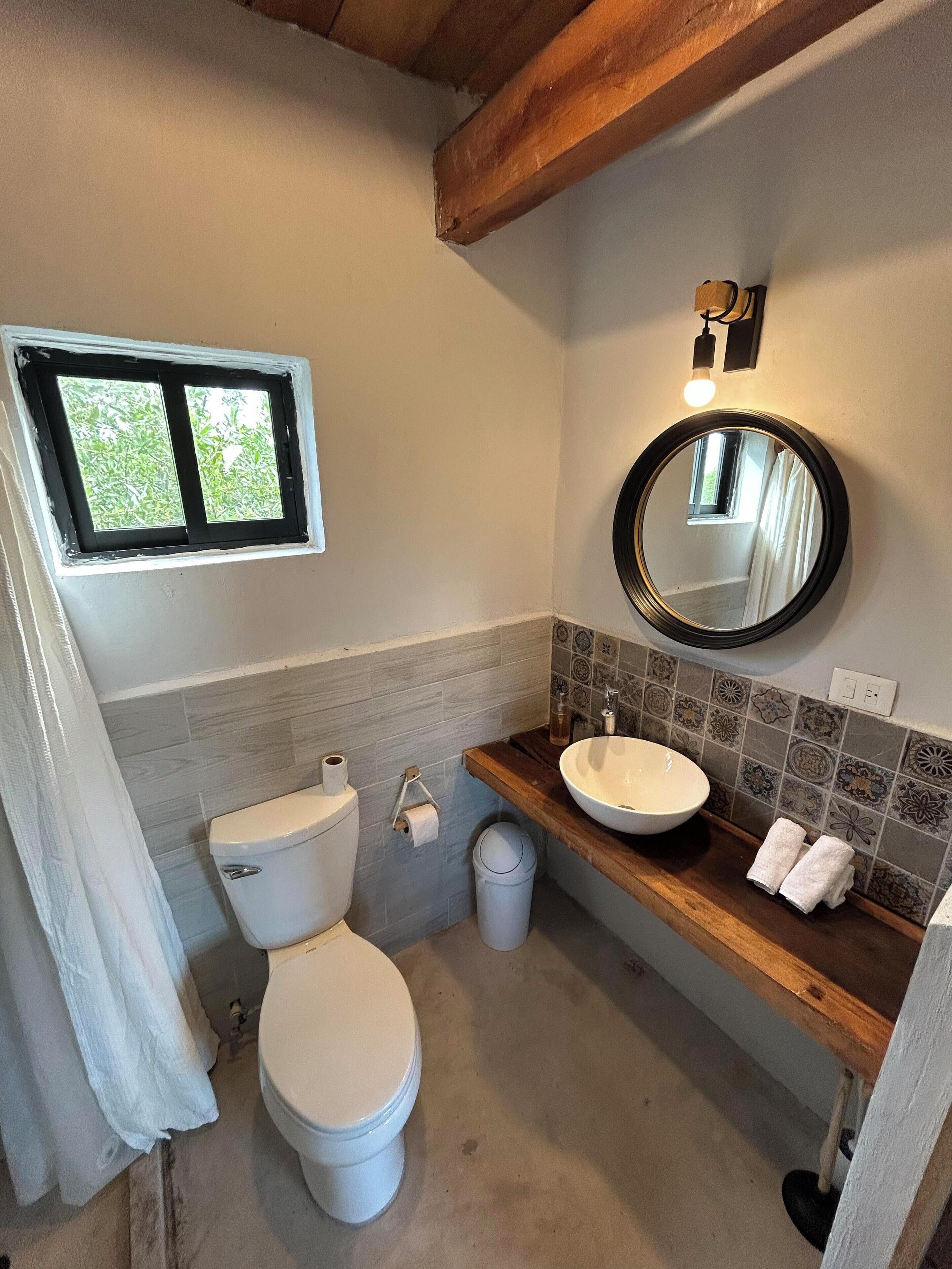 A bathroom with a toilet, sink, mirror, and a window.