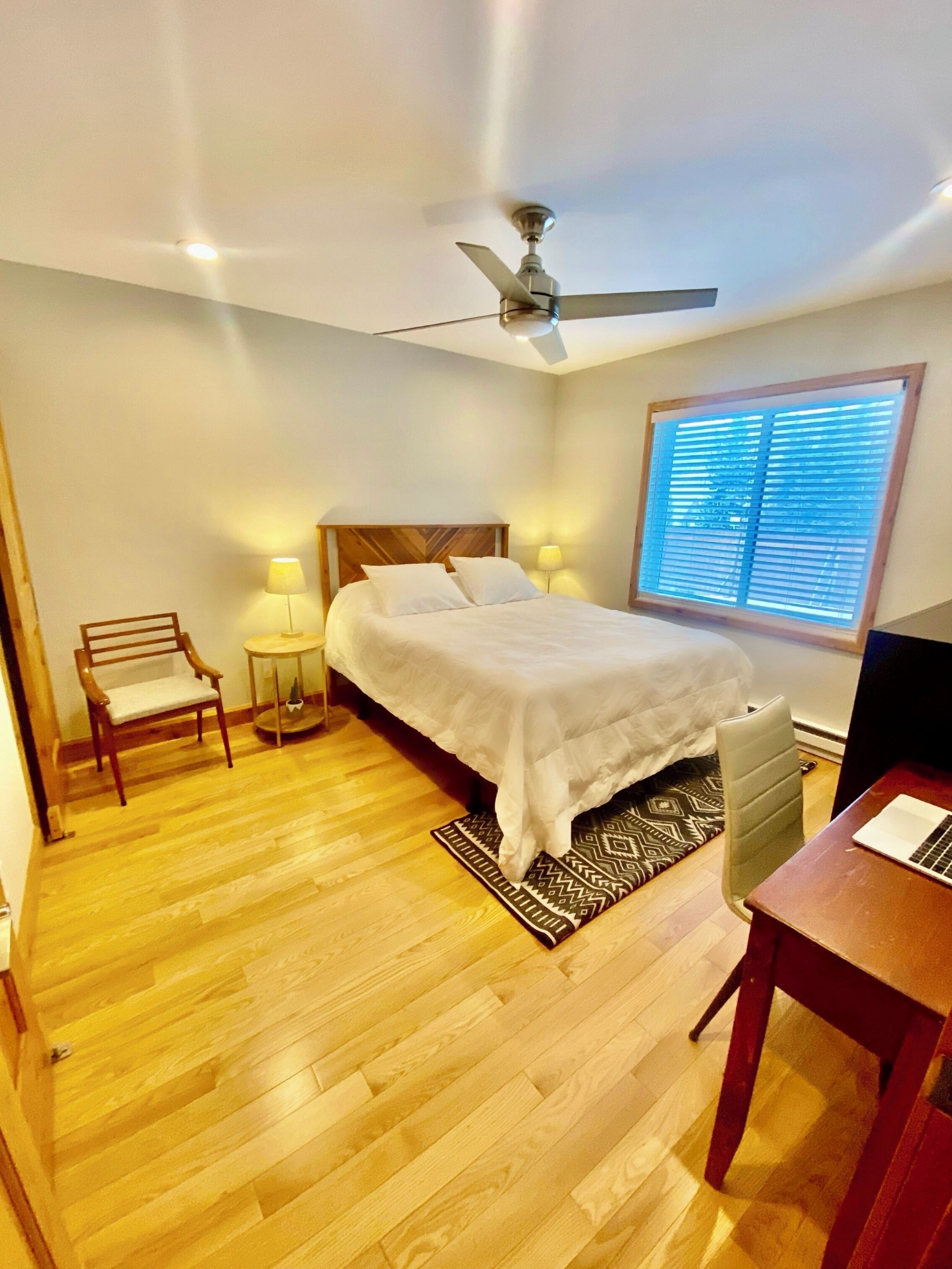 Une chambre à coucher comprenant un lit, un bureau, une chaise, un ventilateur de plafond, une fenêtre avec des stores et un plancher en bois.
